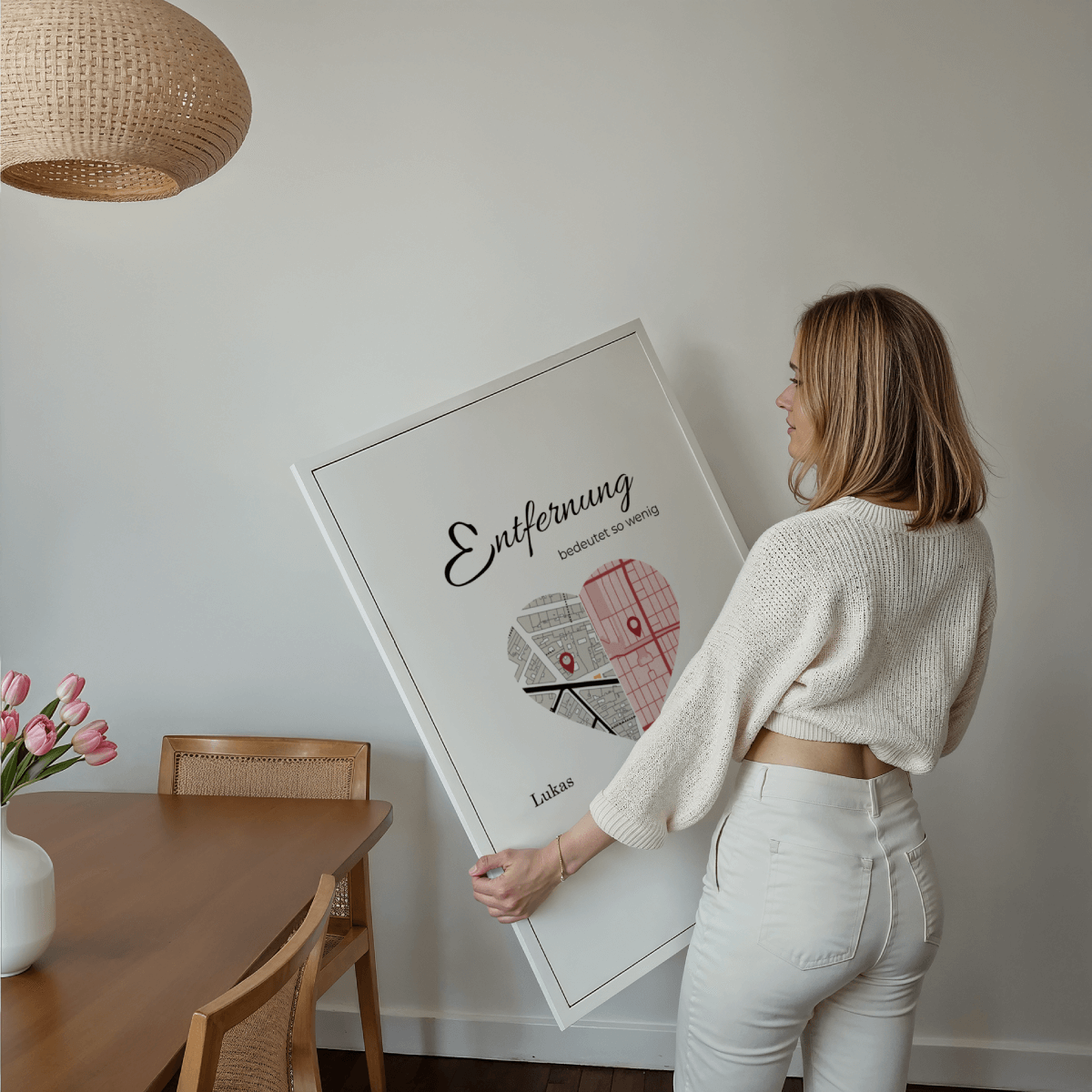Junge Frau hält ein großes, eingerahmtes Poster mit personalisierter Herzkarte und dem Titel 'Entfernung bedeutet so wenig' in einem modernen Esszimmer. Geschenkidee für Fernbeziehung, Freundschaft oder Familie.